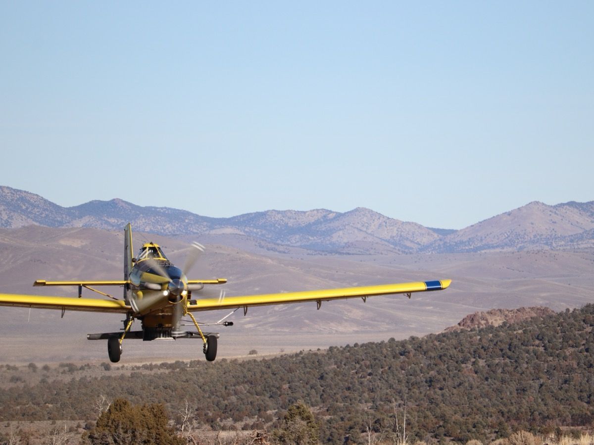 Yellow crop-duster airplane flying low over a desert valley with mountains in the background