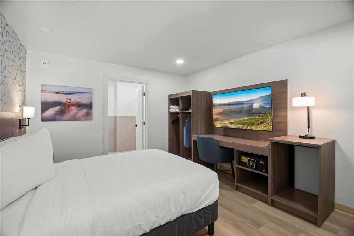 Modern hotel room with white bed, wall art, desk, chair, lamp, and TV stand