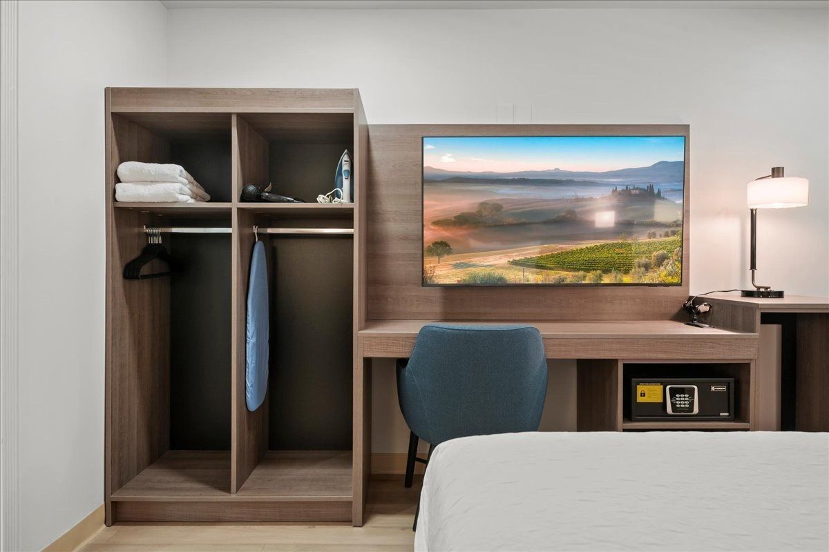 Modern hotel room with TV, desk, chair, open wardrobe, and bed corner.
