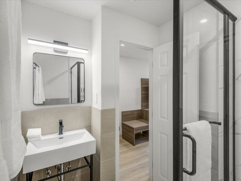 Modern bathroom with white sink, mirror, and open door to a bedroom with wood furniture.