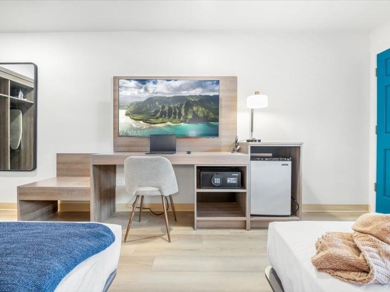 Modern hotel room with a desk, chair, lamp, mini fridge, blue door, and beds visible in the foreground