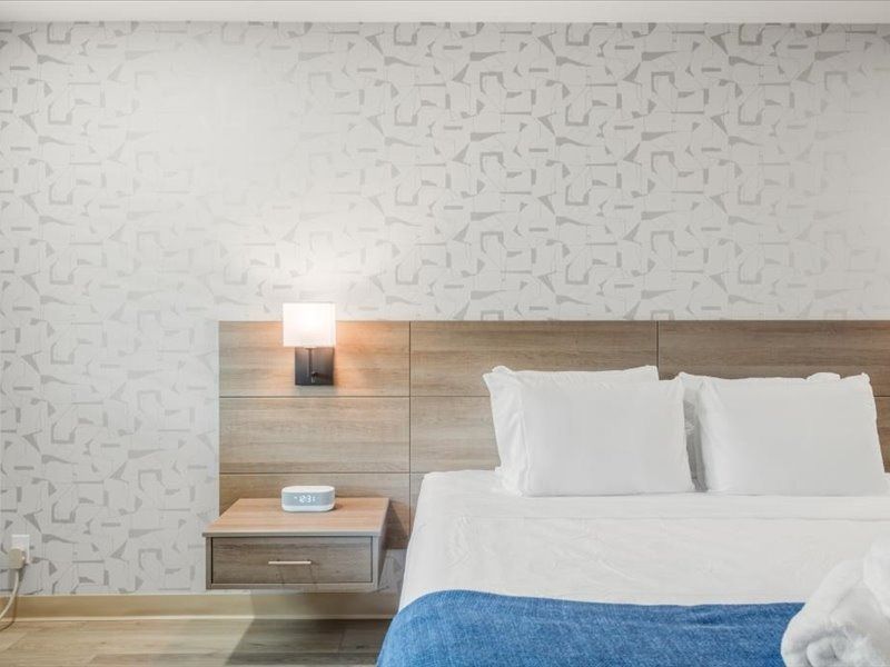 Modern bedroom with a wooden bed, white bedding, bedside lamp, and patterned wallpaper.