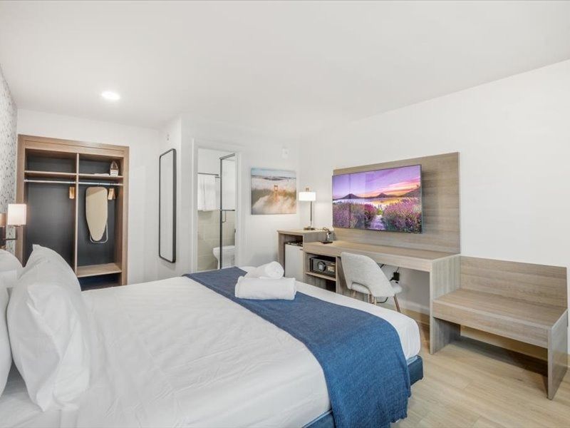 Bright hotel room with queen bed, desk, TV, wardrobe, and bench in a modern white interior