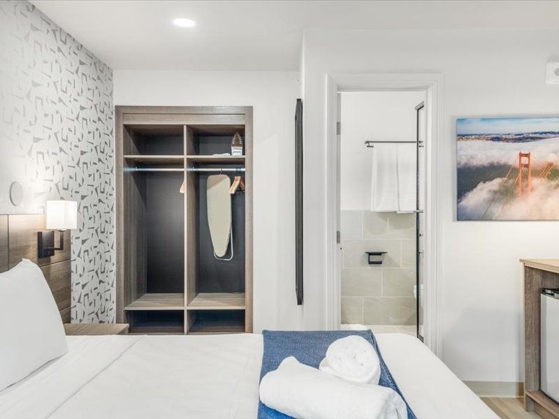 Modern hotel room with open closet, white bed, blue runner, and bathroom visible through an open door