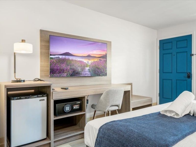 Modern motel room with twin beds, blue door, desk, chair, mini fridge, and wall-mounted TV