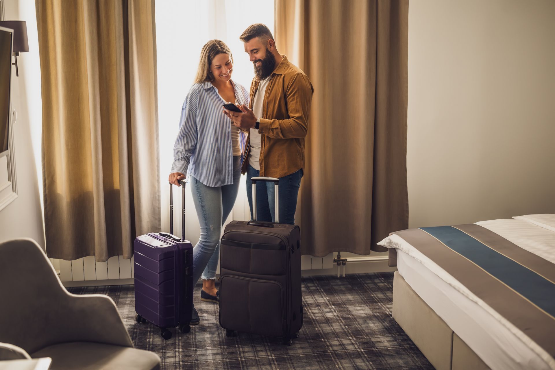 Two people packing in a hotel room beside suitcases and a bed, looking at a phone.