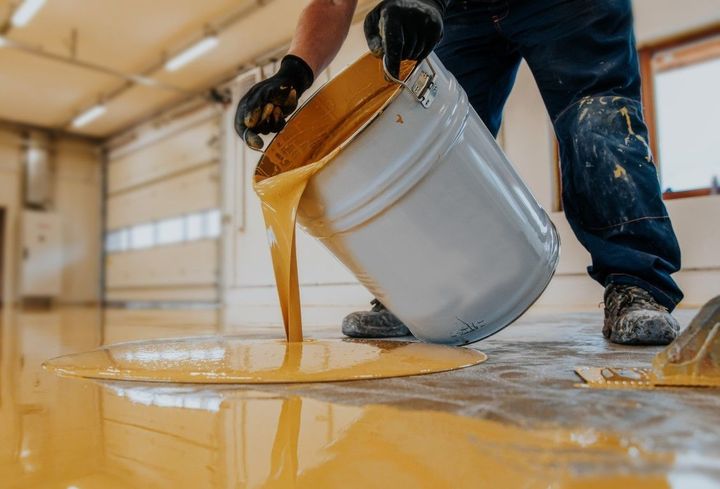 Person pouring yellow paint from a white bucket onto a floor indoors