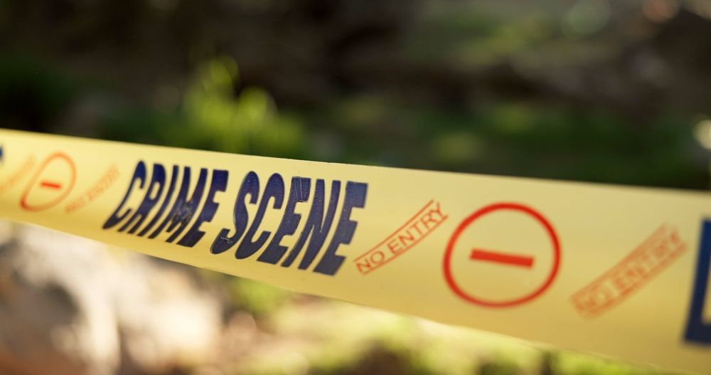 Yellow crime scene tape stretched across an outdoor area, with blurred greenery in the background