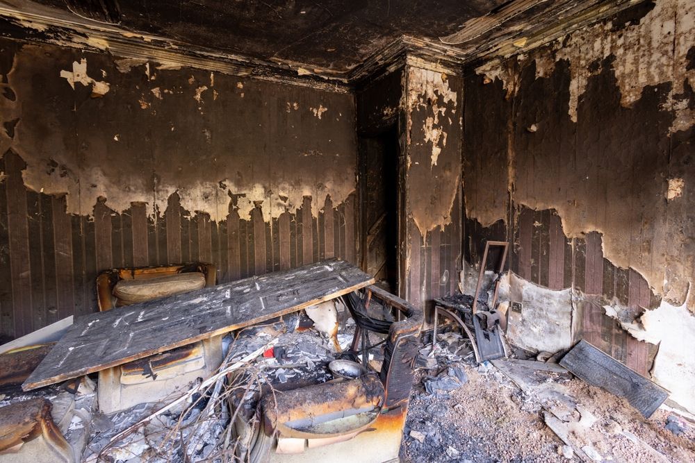 Burned room with charred walls, collapsed debris, and a damaged bed or bench near the back wall