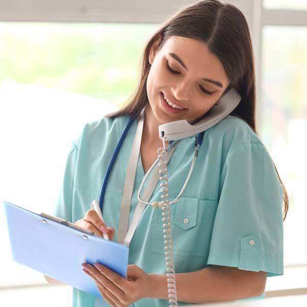 A smiling healthcare worker in a teal uniform, using a phone while taking notes on a blue clipboard. Contact Victory Medical Institute to Discover The Personal PMDD Treatment Plan That Will Work For You