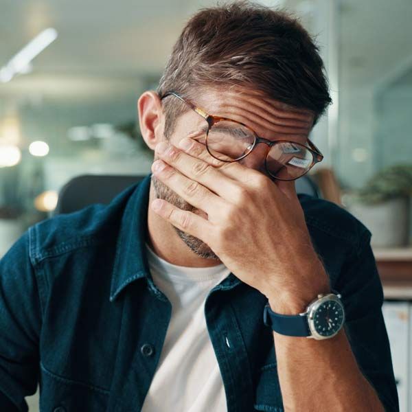 Man with Adrenal Fatigue in glasses, rubbing his face at a desk, looking stressed, with blurred office background