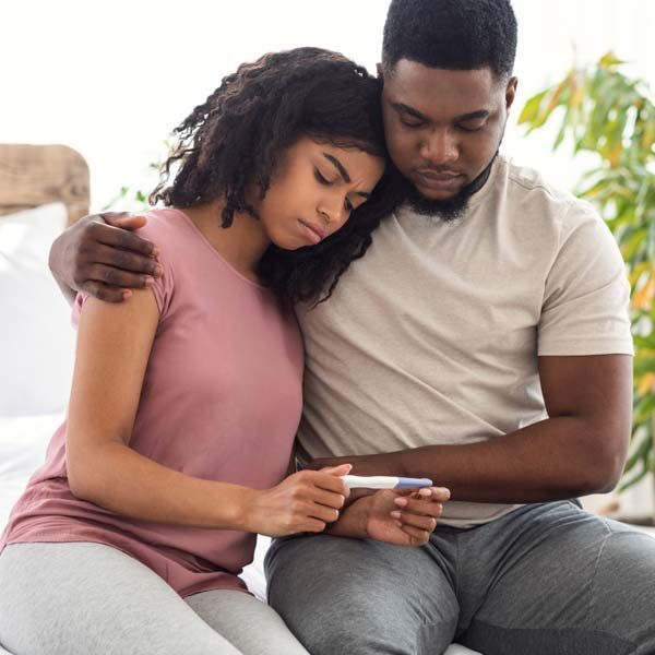 Two people with infertility are sitting on a couch, leaning together and looking at a pregnancy tester with a sad expression.