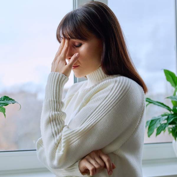 A woman with Low Estrogen in a white sweater standing by a window, pinching her nose with a moody expression.