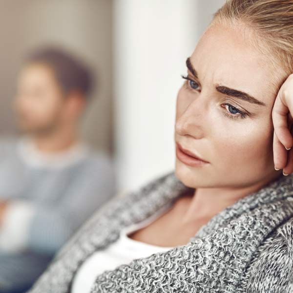 Woman with DHEA Imbalances in gray sweater looking moody while leaning her head on her hand, with a blurred man in the background