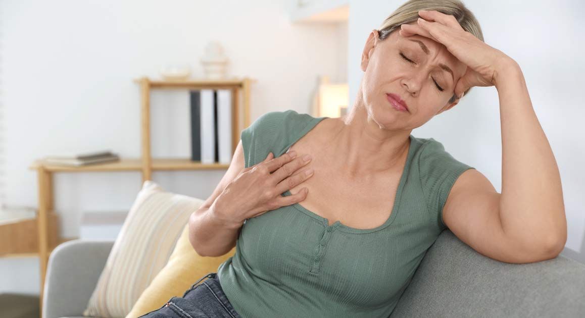 Woman with hot flashes waiting for Menopause Treatments by Dr. Kamal Kalsi, sitting on a couch, holding her forehead and chest, looking unwell in a bright living room