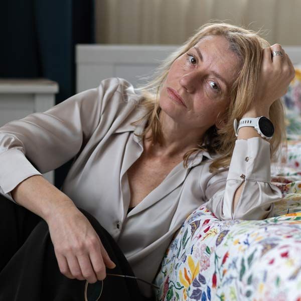Woman with Perimenopause reclining on a bed, resting her head on her hand, wearing a gray blouse and a white watch.