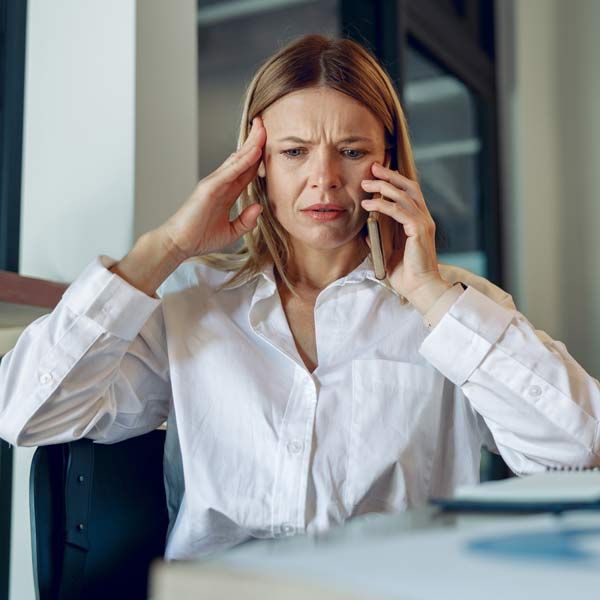 Woman with Perimeopause in a white shirt holding her head and phone, looking stressed at a desk in an office