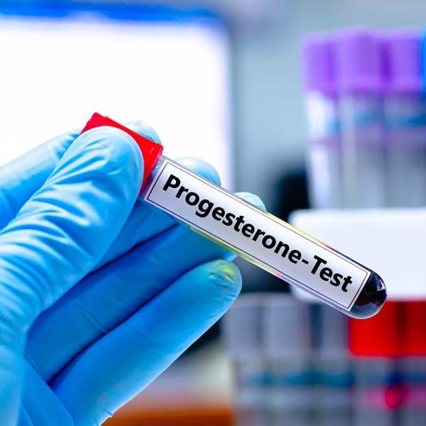Gloved hand holding a lab test tube labeled “Progesterone-Test” with blurred vials in the background