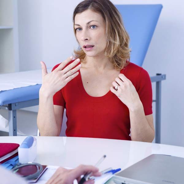 Woman in a red shirt speaking in a medical exam room, explaining her menopause symptoms to Dr Kamal Kalsi