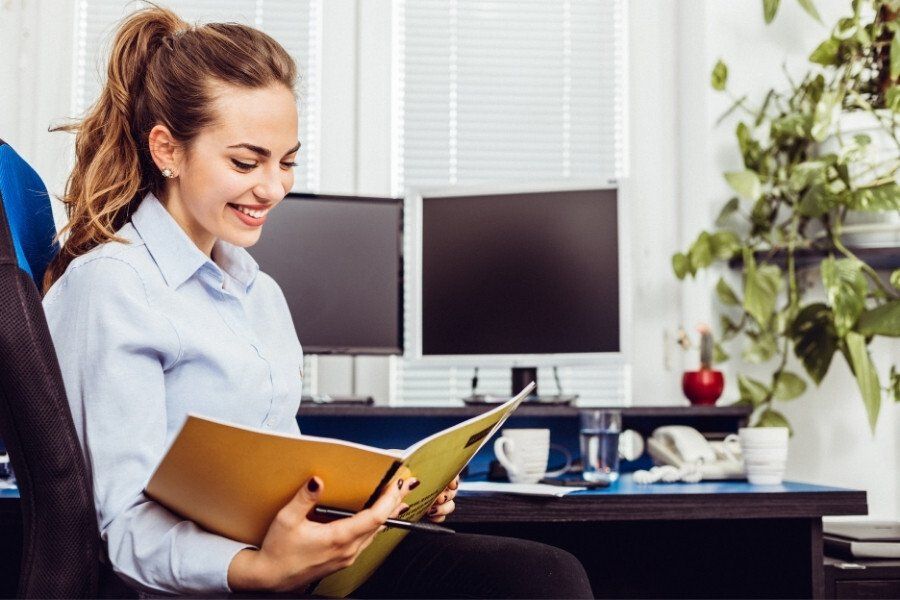 Woman smiling while reviewing papers in an office, with two computer monitors and a desk in the background.