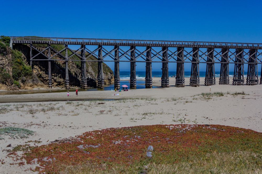 Wooden trestle bridge over a sandy beach with ocean in the background under a clear blue sky