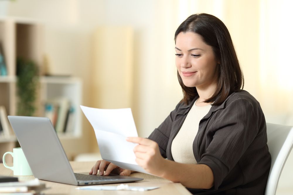 Woman seated at a desk, looking at a document, laptop in front of her.