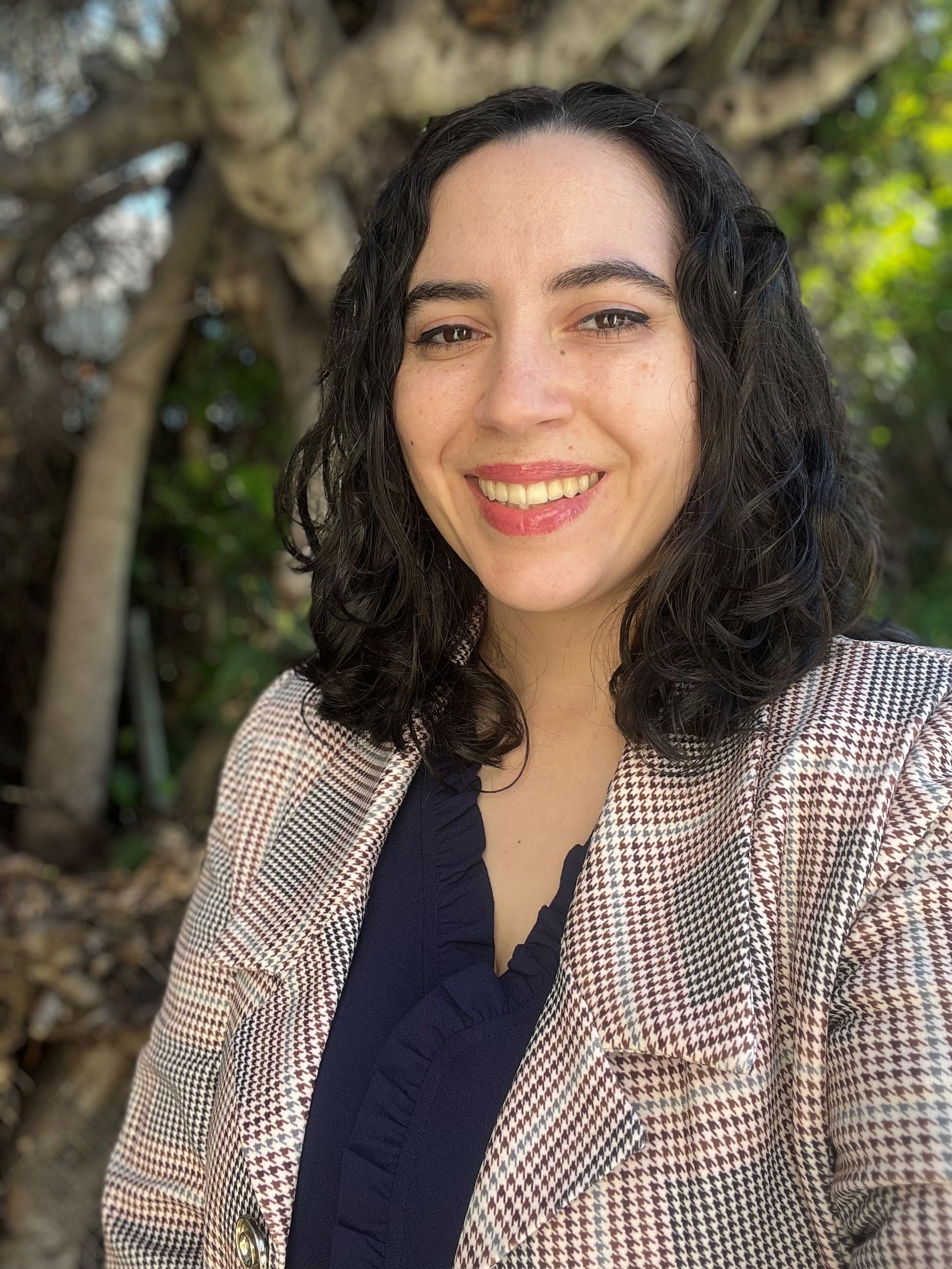 Woman with curly dark hair smiles outdoors, wearing a patterned blazer over a dark blue top.