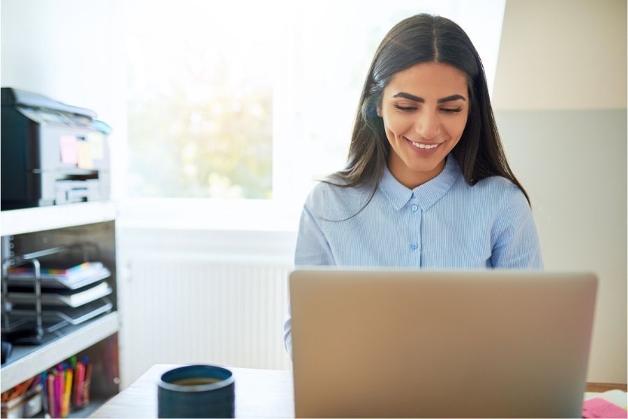 Woman smiles while working on a laptop at a desk near a window; printer and supplies visible.