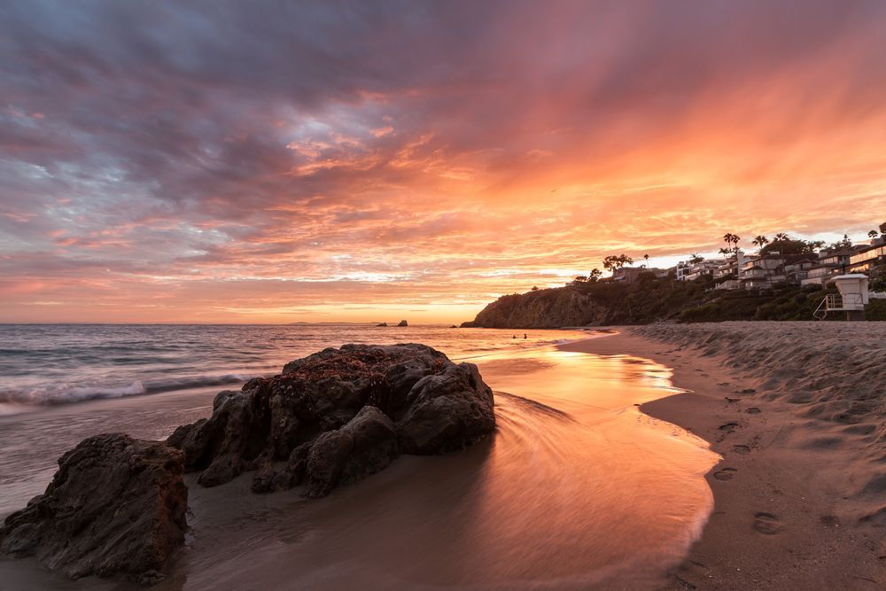 Sunset over a rocky beach with waves, glowing orange sky, and houses on the distant cliff