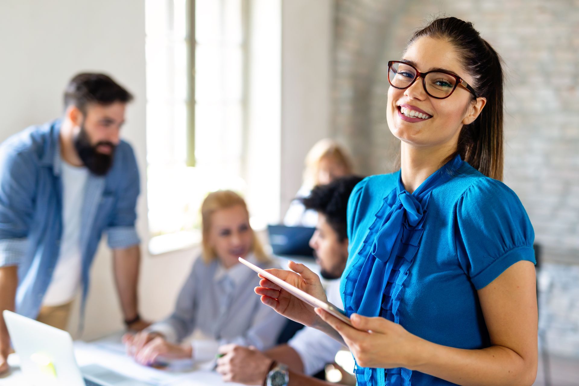 Smiling woman in blue blouse holding a tablet in a bright office meeting with colleagues in background