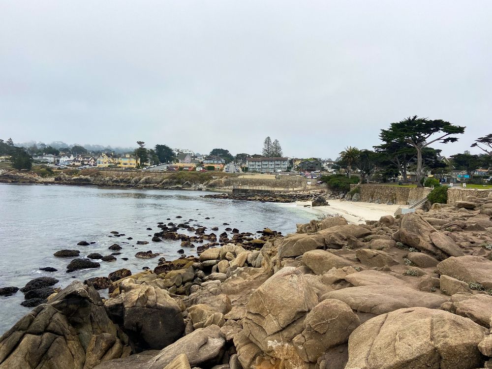 Rocky shoreline with calm water, scattered boulders, and a distant coastal village under an overcast sky