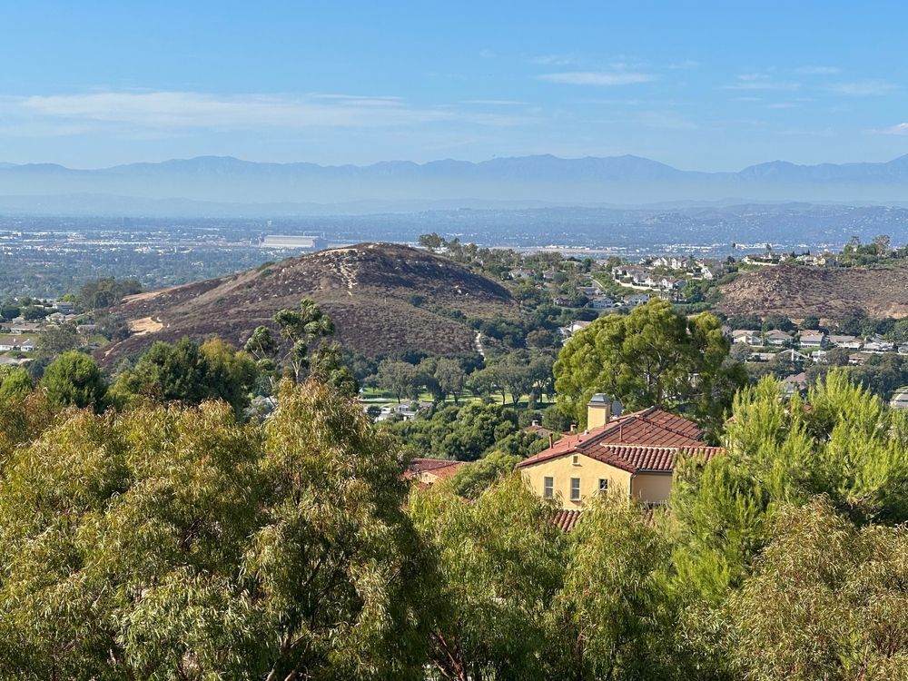 Hazy hillside view over suburban homes and trees toward distant mountains under a blue sky