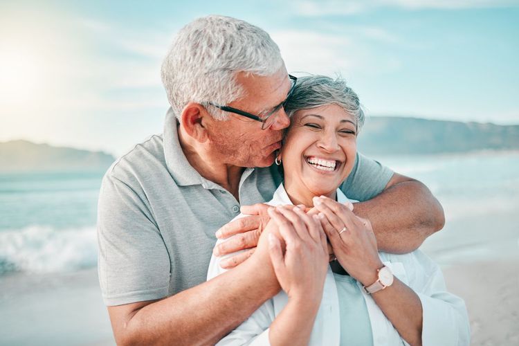 Two smiling people embracing on a beach, with one kissing the other on the head at sunset.