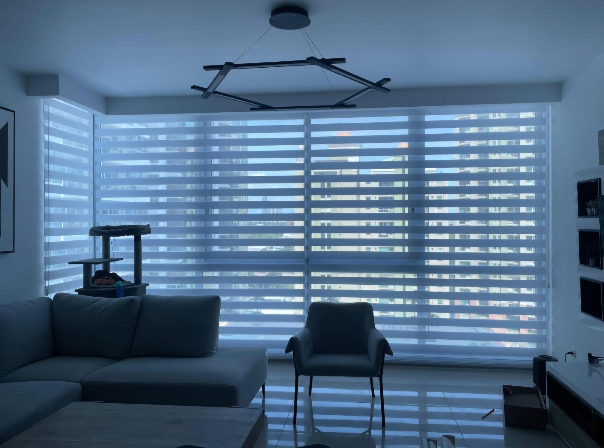 Una sala de estar con un sofá modular gris, un sillón y una gran ventana cubierta por persianas horizontales translúcidas.