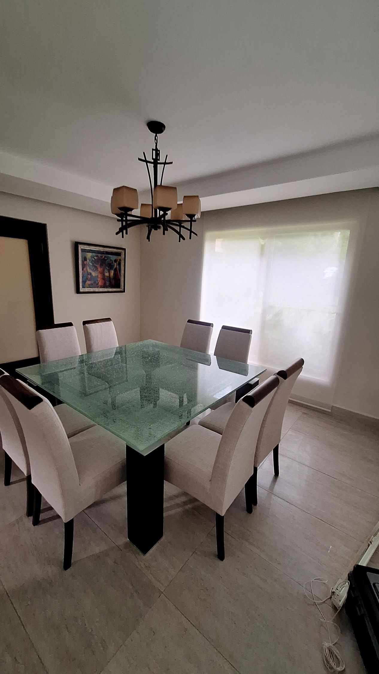 Una mesa de comedor cuadrada de cristal con seis sillas tapizadas en color beige, situada bajo una lámpara de araña