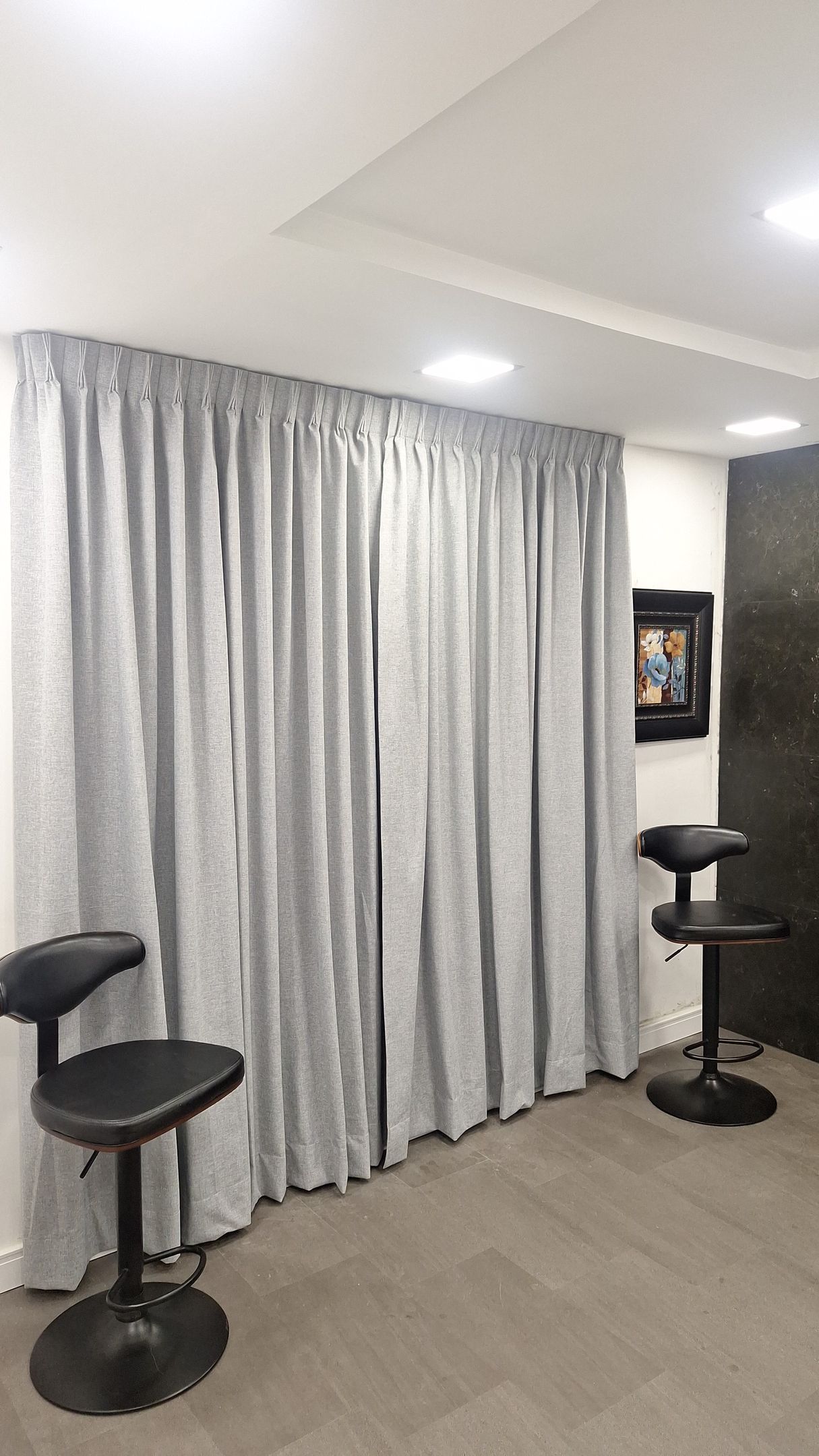 Habitación con cortinas grises, dos taburetes negros y un cuadro en una pared oscura.