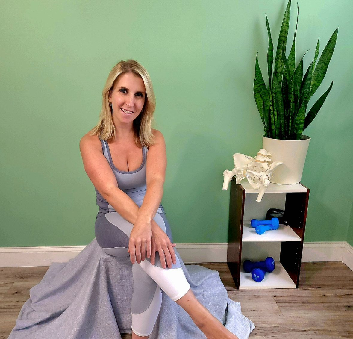 Woman seated on the floor stretching beside a shelf and potted plant in a room with a green wall.