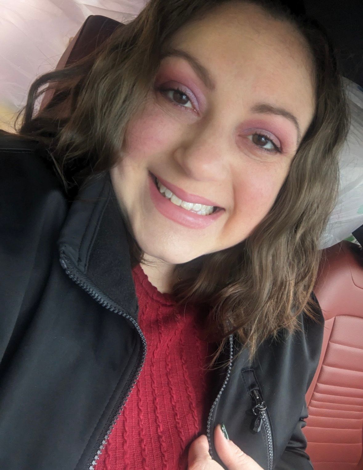 Smiling person in a black jacket and red sweater, seated in a car