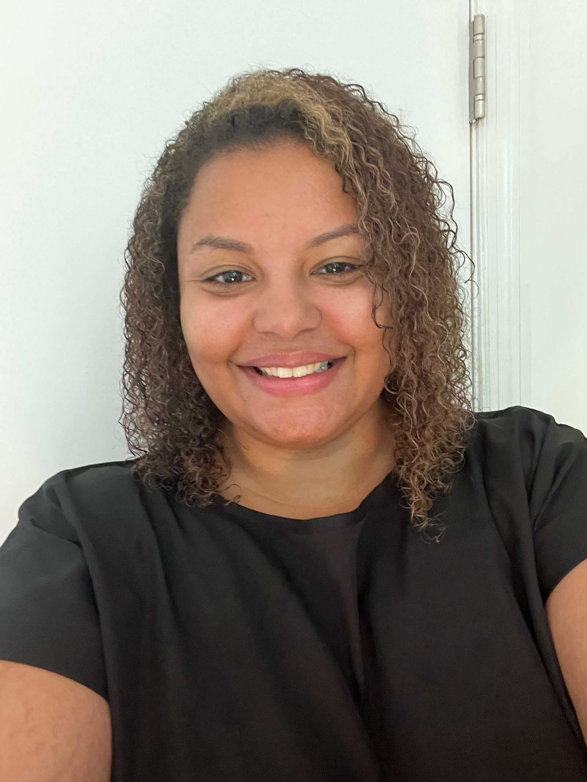 Smiling person with curly hair wearing a black top against a white wall