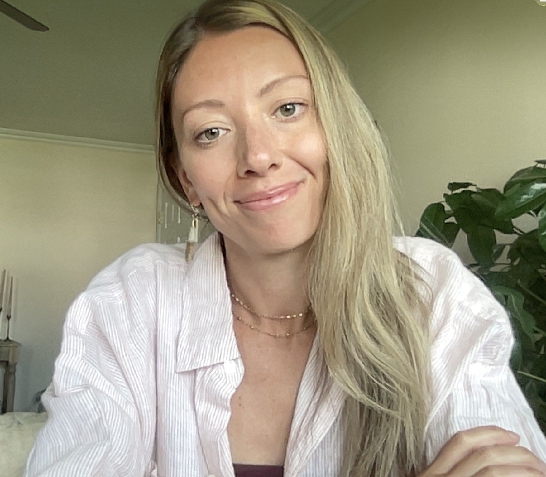 Smiling person with long blonde hair in a white shirt, sitting indoors near a plant.