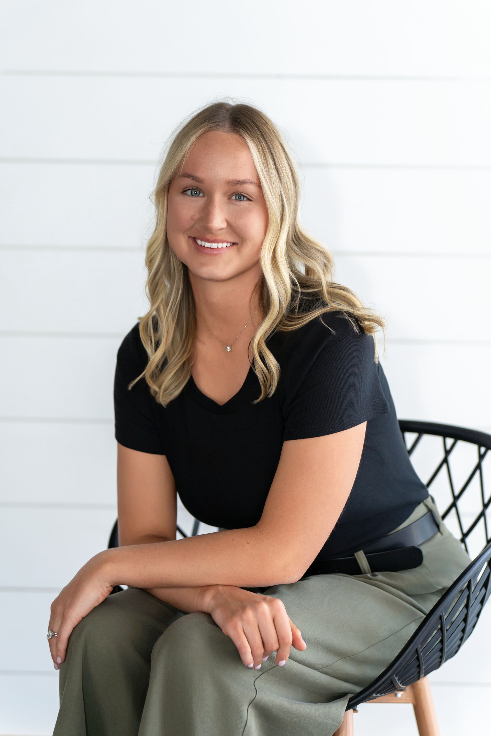 Smiling woman in a black shirt and olive pants seated on a black chair against a white wall