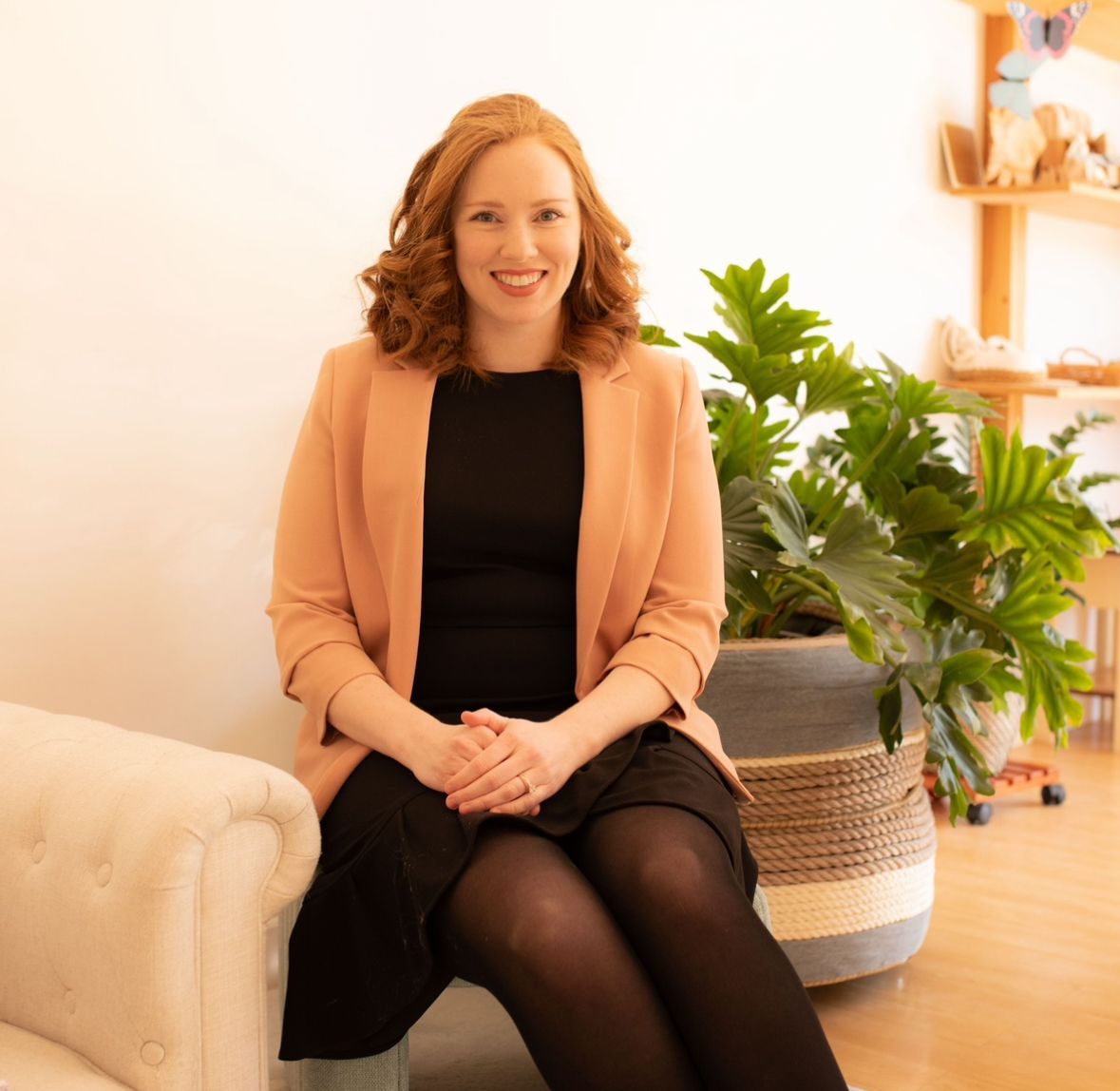 Woman seated in a bright living room beside a large potted plant, smiling at the camera