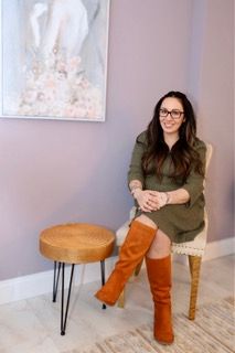 Woman seated beside a small round table in a pastel room, smiling with one leg crossed.