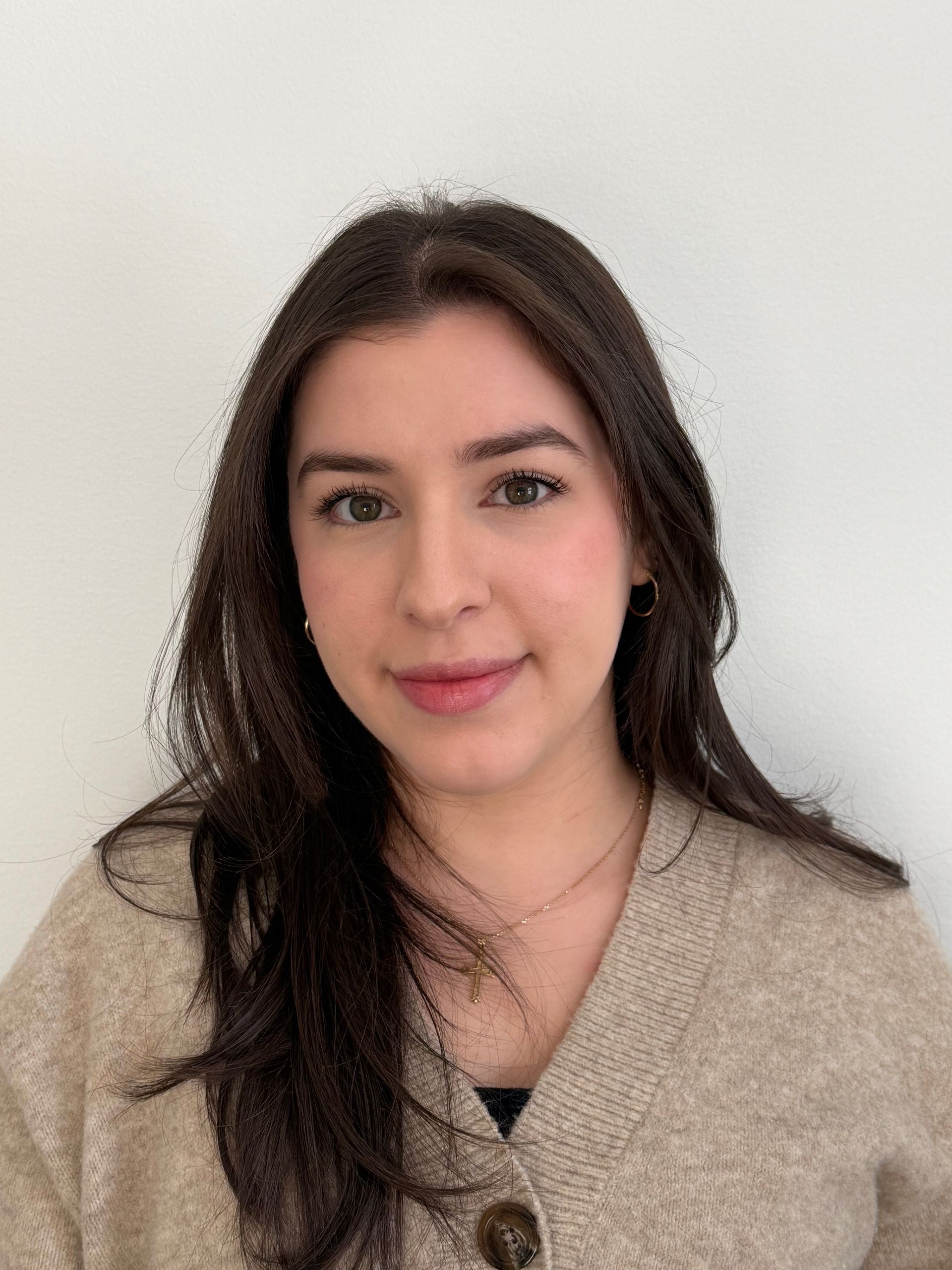 Portrait of a smiling person with long dark hair wearing a beige sweater against a light wall