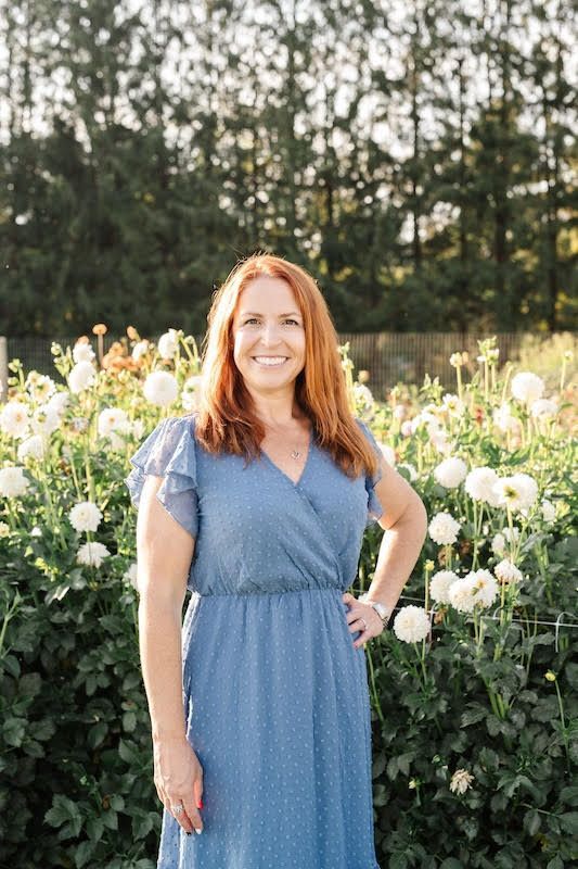 Smiling woman in a blue dress standing in a flower garden with tall white blooms and trees behind her
