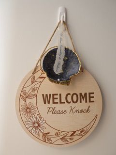 Wooden welcome sign with floral border, saying “WELCOME Please Knock,” hanging on a wall