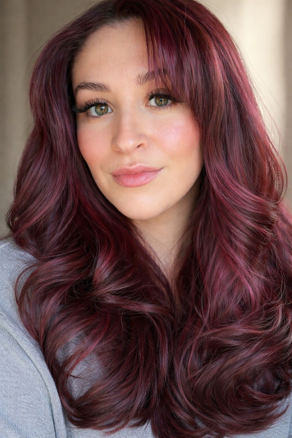 Portrait of a person with long wavy burgundy hair, soft makeup, and a light gray top against a neutral background
