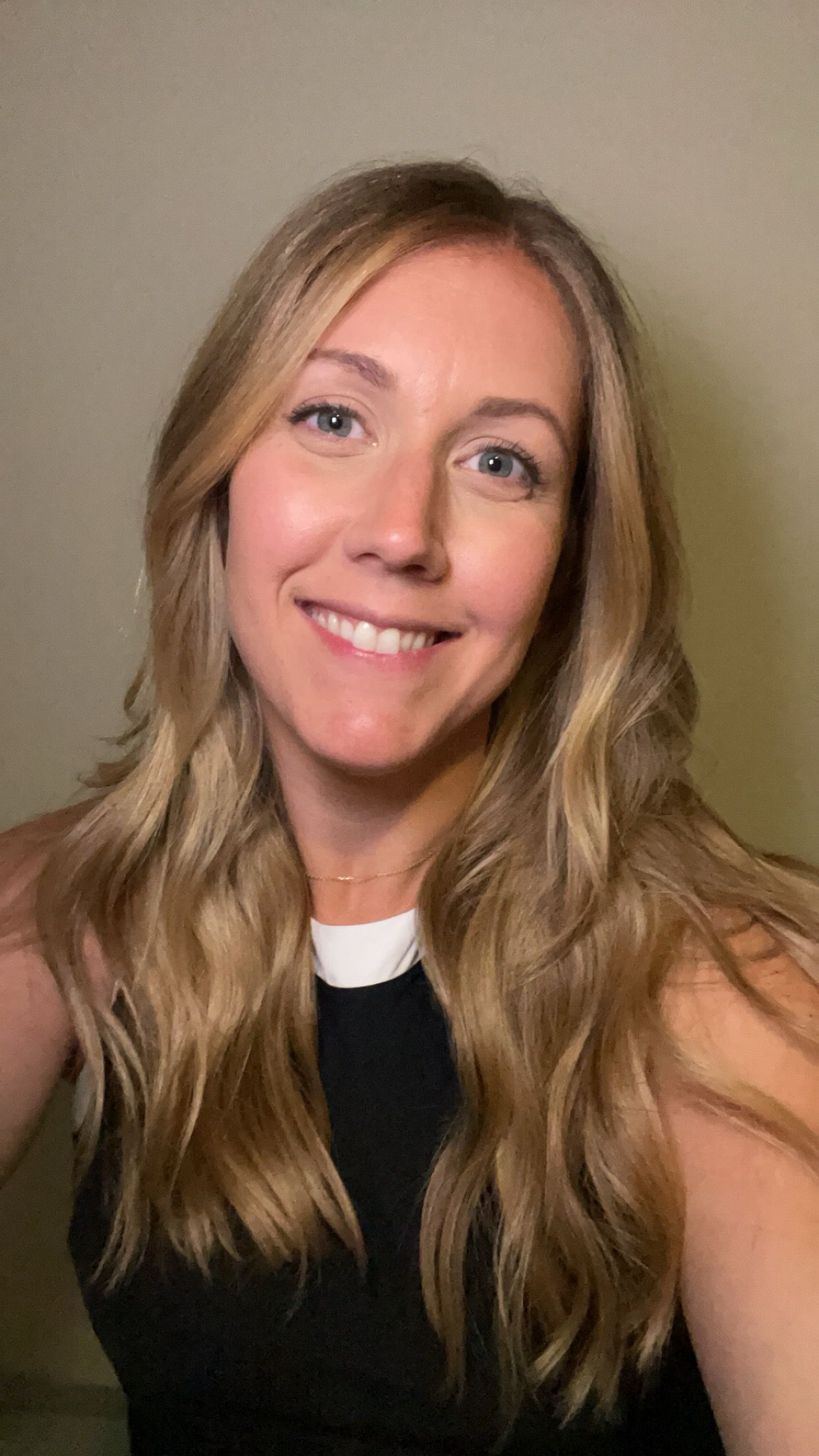 Smiling blonde woman in a black sleeveless top against a plain beige background
