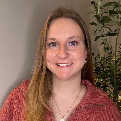 Smiling woman in a pink sweater standing indoors beside a leafy plant.