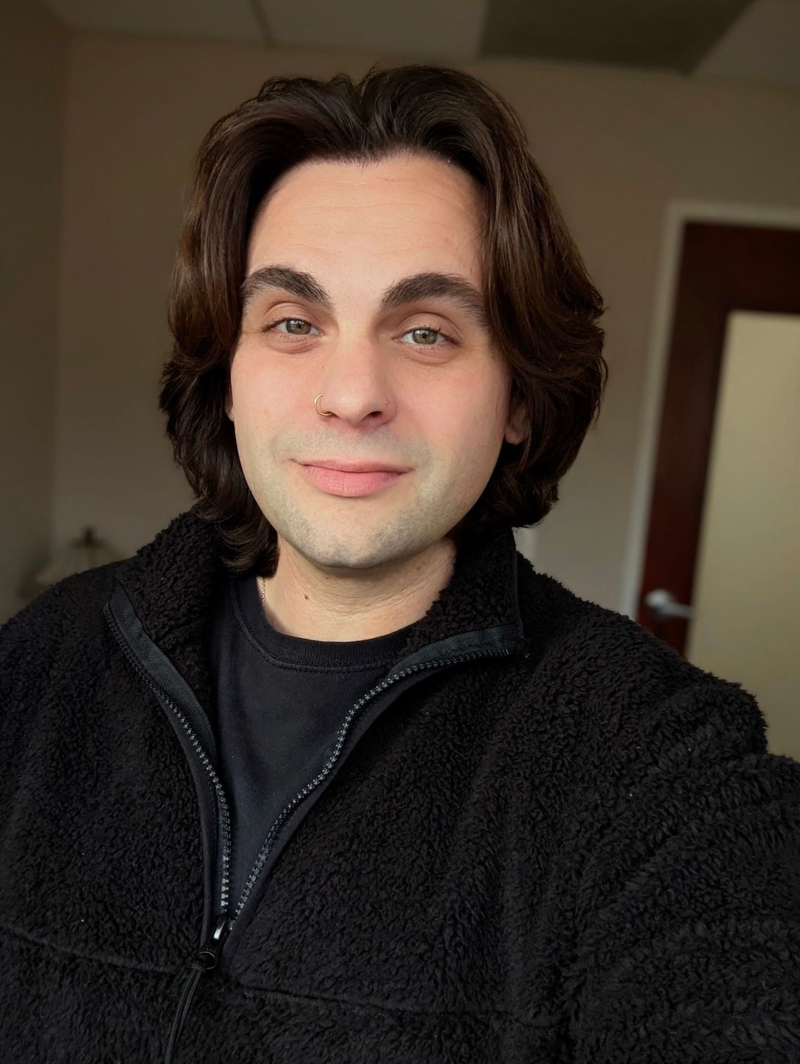 Person with shoulder-length dark hair wearing a black fleece jacket, smiling in a softly lit indoor setting.